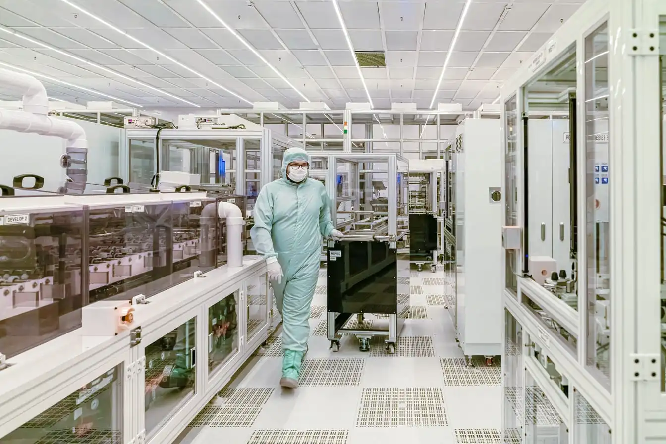 A worker in a full cleanroom suit and mask pushes a cart down an aisle between semiconductor process tools.