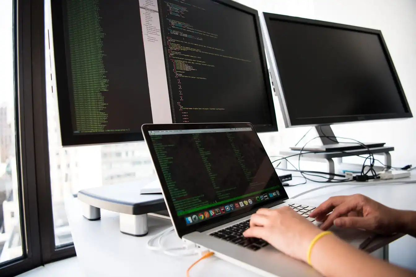 Hands typing on a laptop showing lines of code, with two external monitors on the desk behind it.