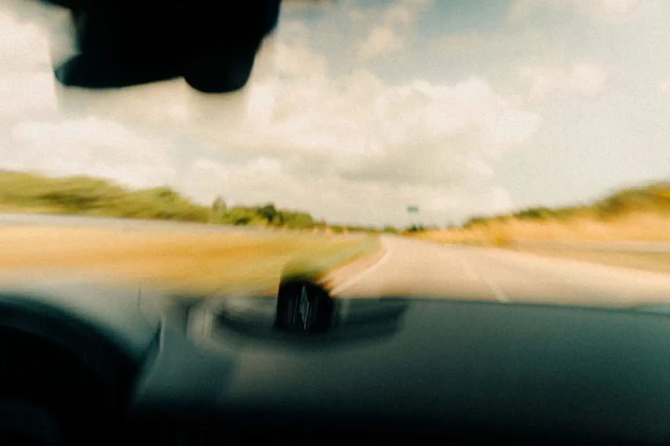 The view forward through a moving car's windshield onto an open road, the roadside blurred by speed.